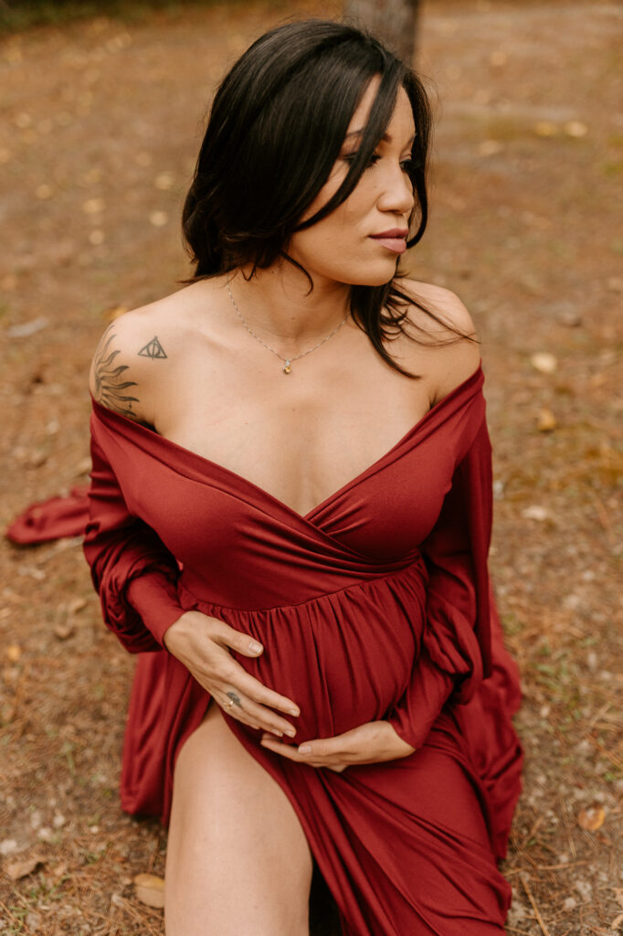 Pregnant woman kneeling on forest floor looking over her shoulder to the right in a maroon dress with a slit on her right knee while holding her belly, photo taken by professional maternity photographer 